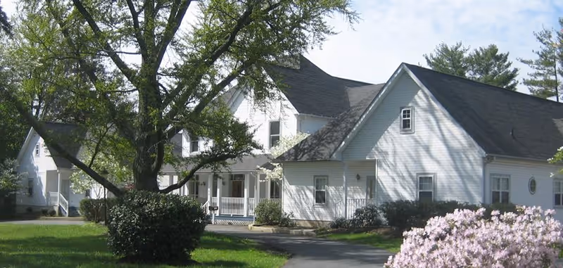 Exterior view of a white senior living facility building with a dark roof, surrounded by green trees, bushes, and blooming flowers under a clear sky.
