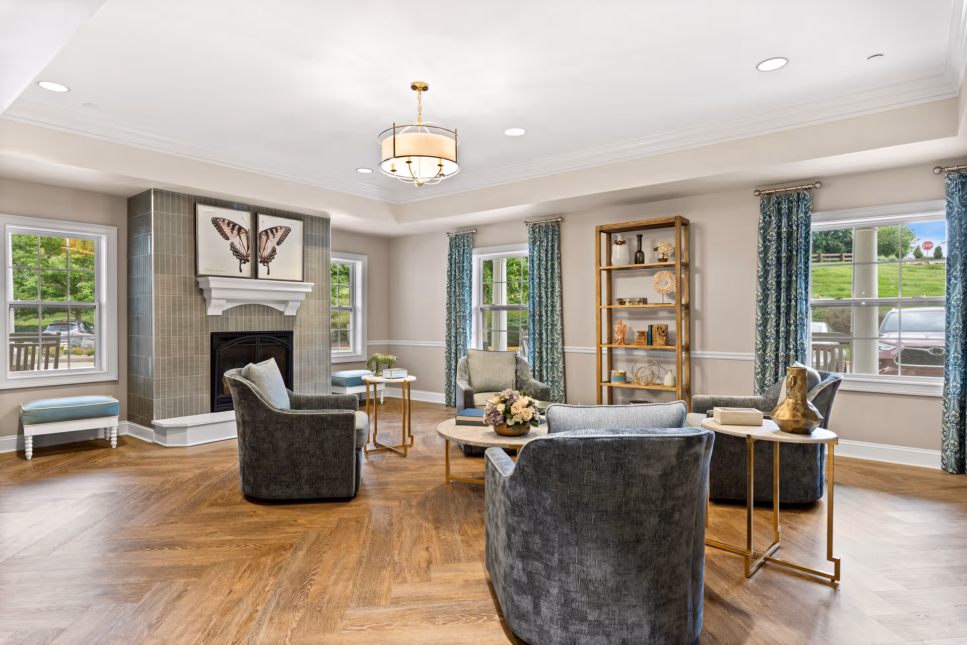 A bright and spacious living room with four gray upholstered armchairs arranged around a round coffee table with a floral centerpiece. The room features a fireplace with a butterfly artwork above it, large windows with blue patterned curtains, a wooden floor, and a tall wooden shelving unit with decorative items.