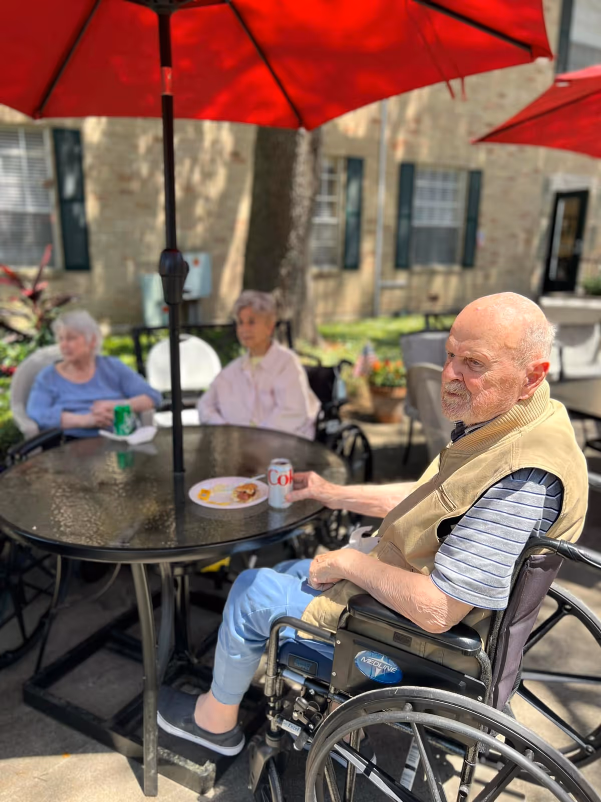 Three elderly residents sit at a round outdoor patio table under red umbrellas, with one man in a wheelchair holding a can of Diet Coke.