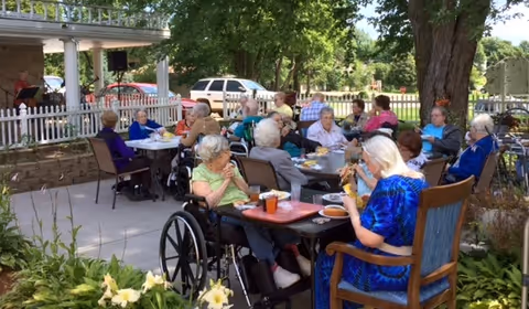 A group of elderly people sitting outdoors at tables enjoying a meal together in a garden patio area with trees and plants around. Some individuals are in wheelchairs, and there is a white picket fence and a parked car in the background.