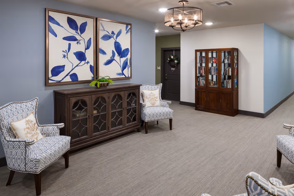 A cozy interior hallway area in a senior living facility featuring two patterned armchairs with decorative pillows, a wooden cabinet with glass doors, and two large framed artworks of blue leaves on the wall. There is a ceiling light fixture and a door with a wreath in the background.