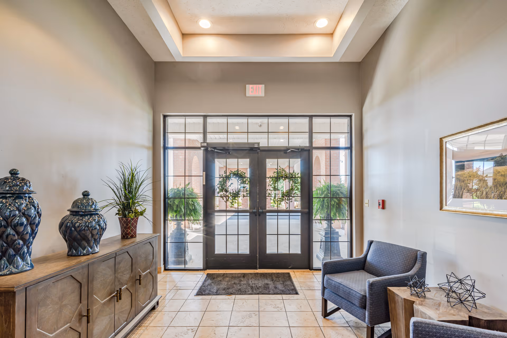 Entrance area of a senior living facility with double glass doors decorated with wreaths, two large potted plants outside, a wooden cabinet with decorative blue vases and a potted plant on the left, and a gray armchair with a wooden side table holding decorative metal sculptures on the right.
