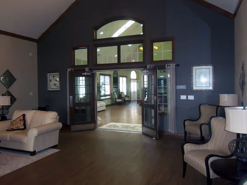 Interior view of a senior living facility lounge area with beige and striped armchairs, a beige sofa, lamps, framed artwork on the walls, and double glass doors leading to a well-lit hallway with additional seating and large windows.