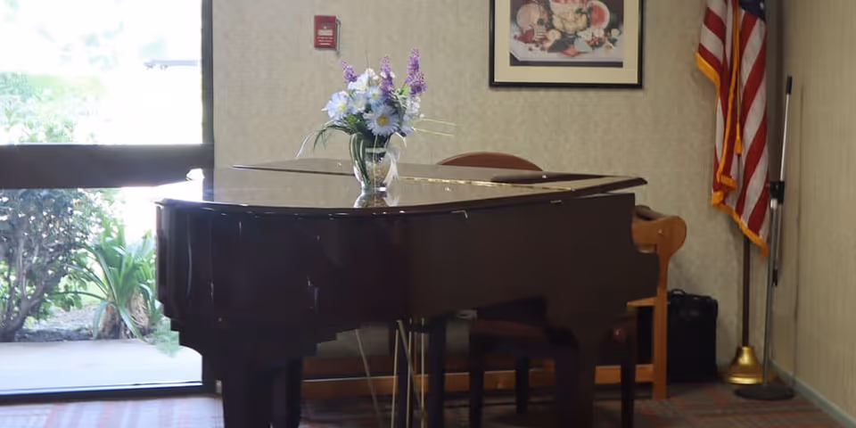 A grand piano with a vase of flowers on top in a lounge area beside a window and an American flag.