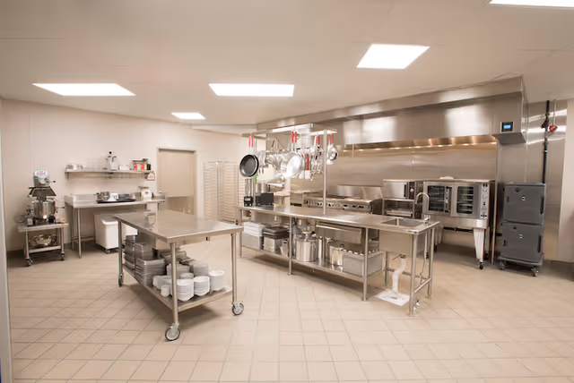 Large clean commercial stainless-steel kitchen with prep tables, hanging pots and pans, ovens, and shelving.