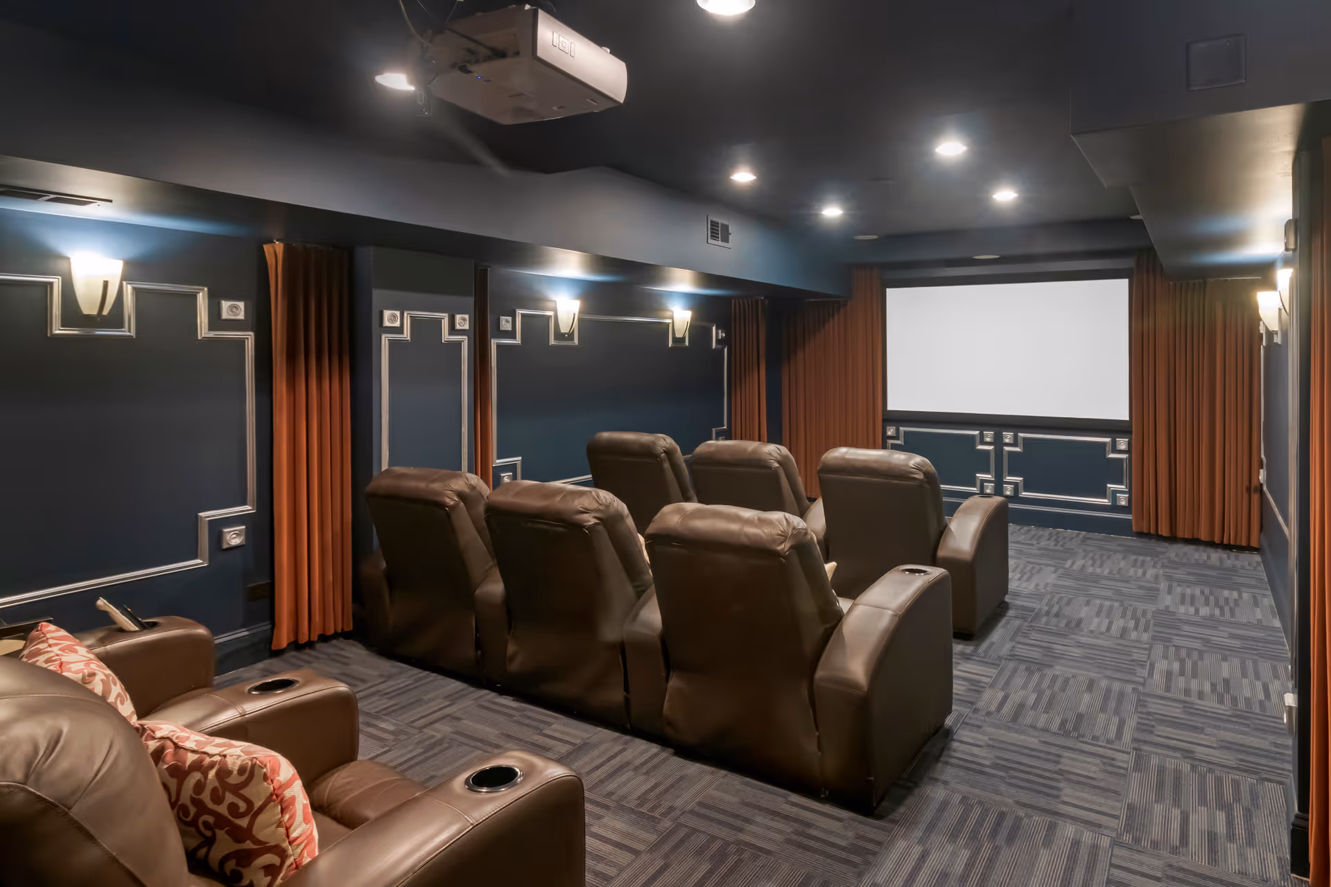 A cozy home theater room with two rows of brown leather recliner chairs facing a large white projection screen. The walls are dark blue with decorative molding and orange curtains on the sides. Ceiling lights and a projector are mounted above.