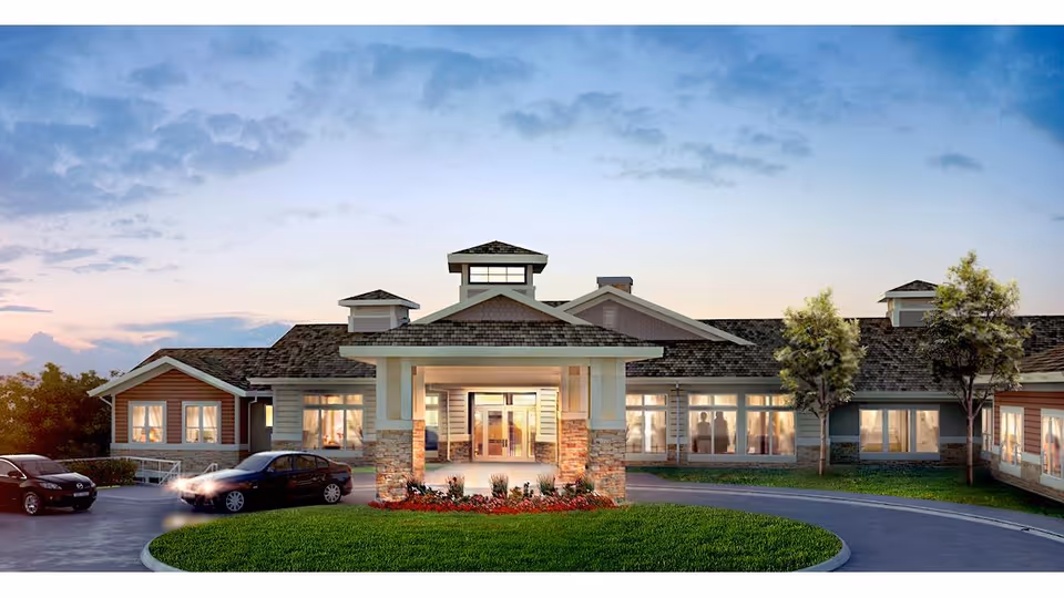 Front exterior of a single-story assisted living facility with a covered entrance, landscaped circular drive, parked cars, and illuminated windows at dusk.