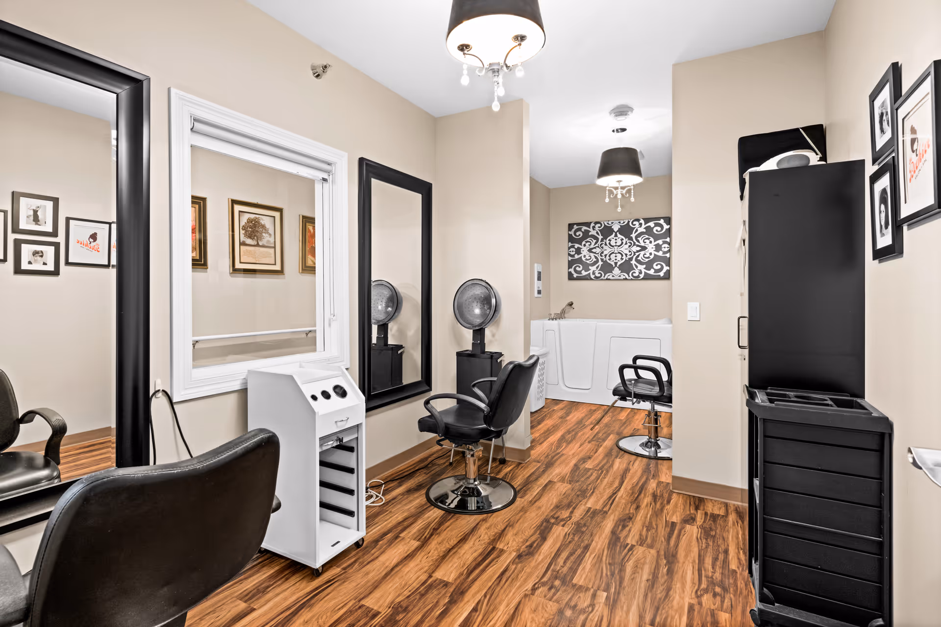Interior view of a hair salon area in an assisted living facility featuring two black salon chairs, large black-framed mirrors, a white hair dryer, a black rolling cart, and a walk-in bathtub in the background. The room has wood flooring, beige walls, framed artwork, and two ceiling light fixtures with black shades.