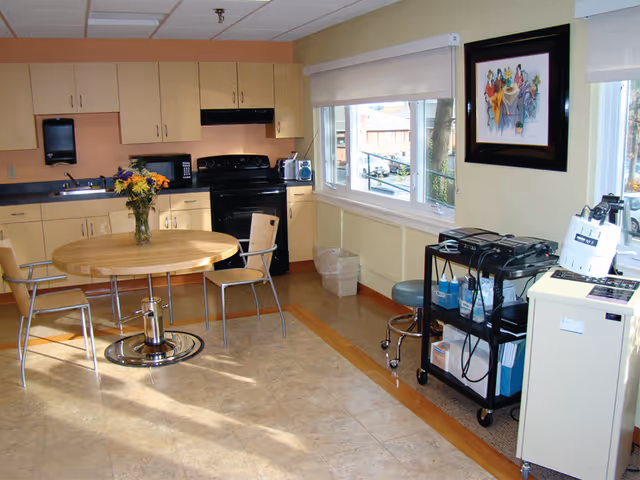 Communal kitchen and dining area with a round wooden table and chairs, light wood cabinets, a black stove, and a window.
