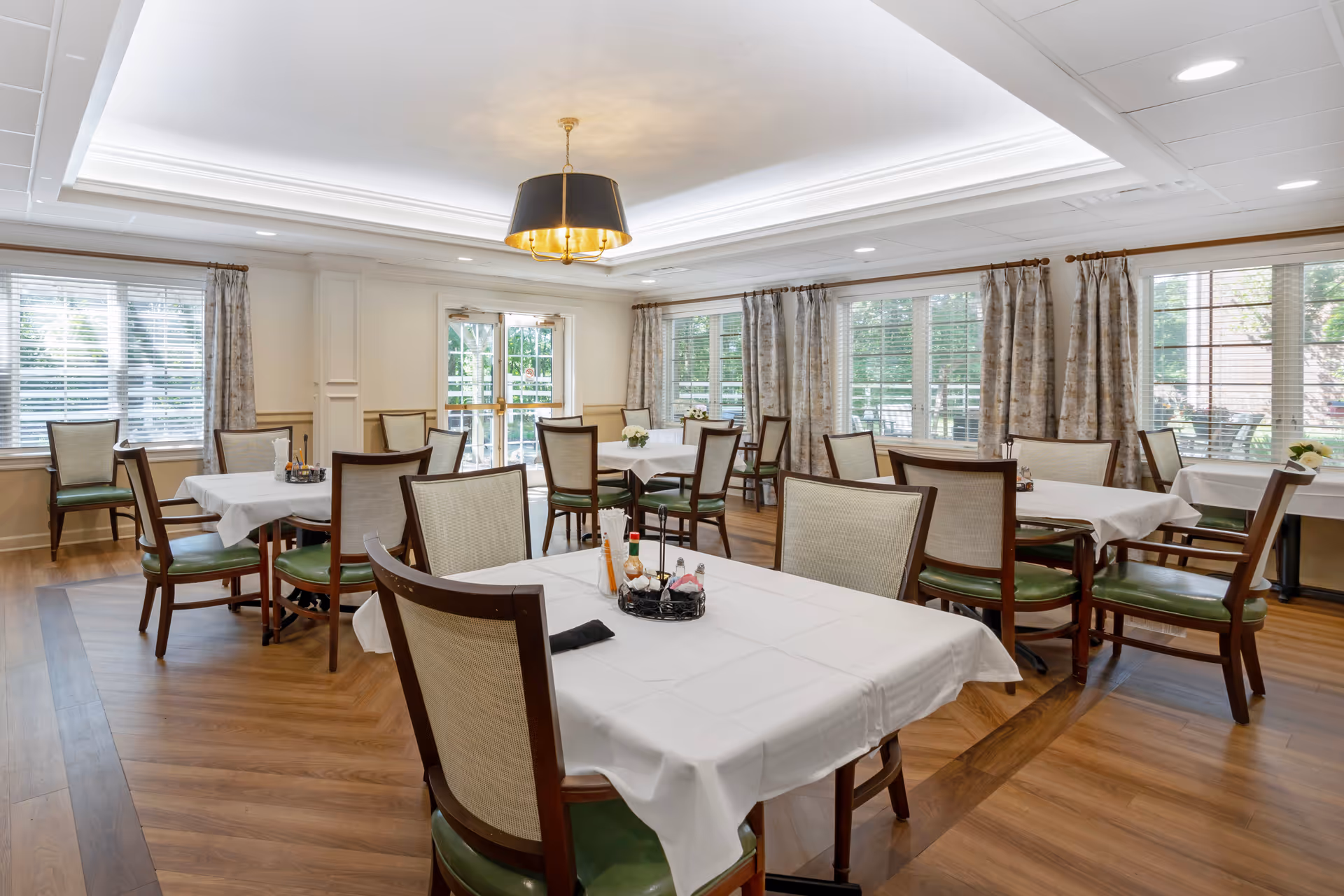 A bright dining room with multiple tables covered in white tablecloths, surrounded by wooden chairs with green cushions. Large windows with curtains allow natural light to fill the room. A modern chandelier hangs from the ceiling, and the floor is made of wood.