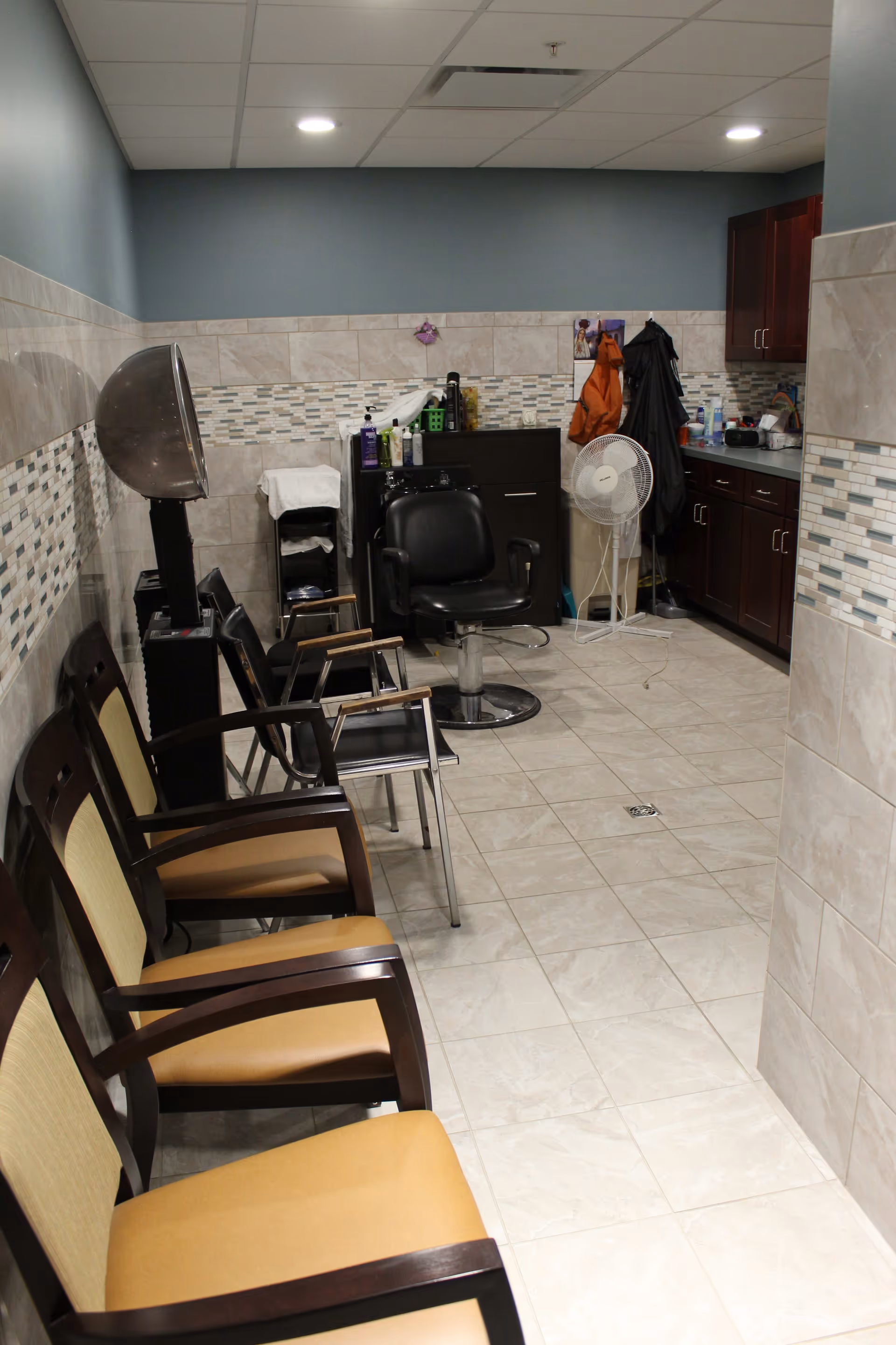 Interior view of a senior living facility hair salon area with a row of waiting chairs on the left, a hair dryer, a black salon chair, cabinets, and various hair care products on the counter and shelves.