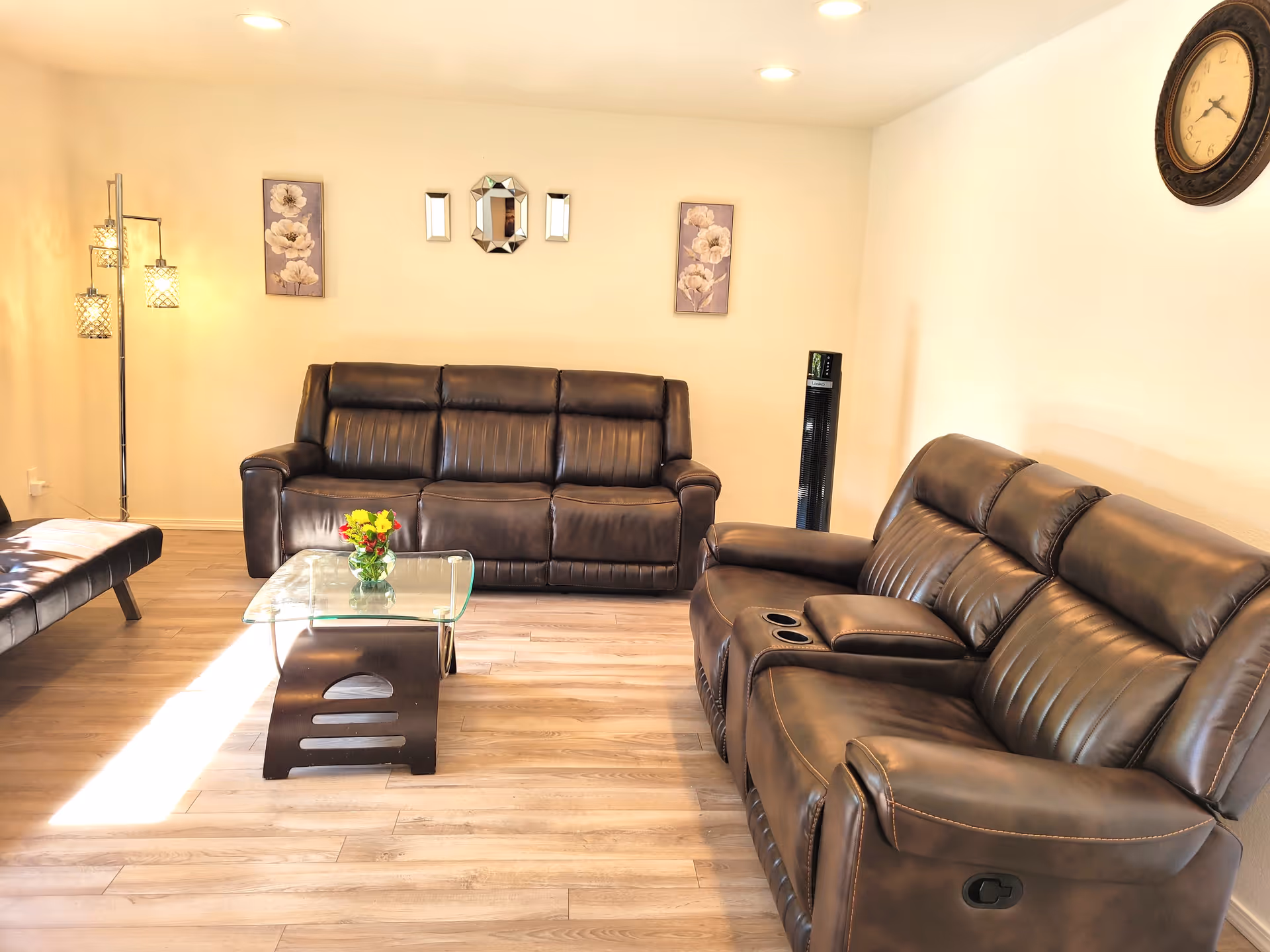 A bright living room with two dark brown leather sofas, a glass coffee table with a small flower arrangement, a standing lamp with three lights, two floral wall paintings, a decorative wall mirror, a tall black fan, and a round wall clock.