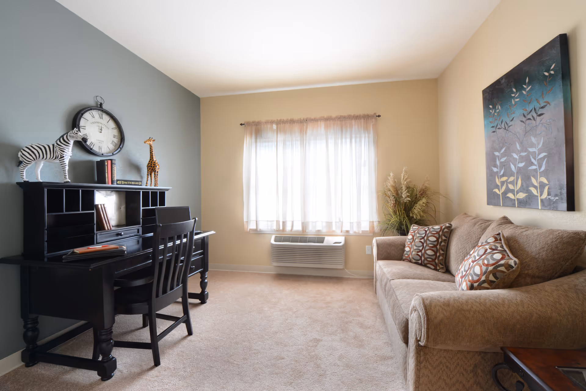 A cozy living room with a beige couch adorned with patterned pillows, a large window with sheer curtains letting in natural light, a black wooden desk with a matching chair, decorative animal figurines, books, and a wall clock above the desk. A piece of artwork featuring leaves hangs on the wall above the couch, and a plant is placed in the corner near the window.