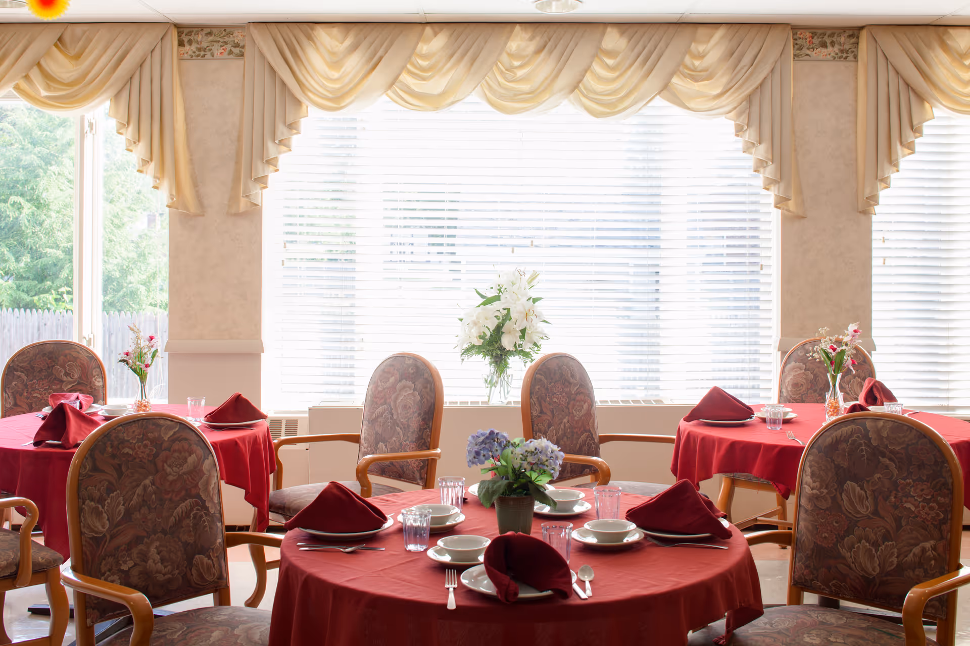A dining room with round tables covered in red tablecloths, each set with plates, bowls, glasses, silverware, and folded red napkins. The chairs have floral upholstery and wooden armrests. There are flower arrangements on the tables and large windows with beige drapes letting in natural light.