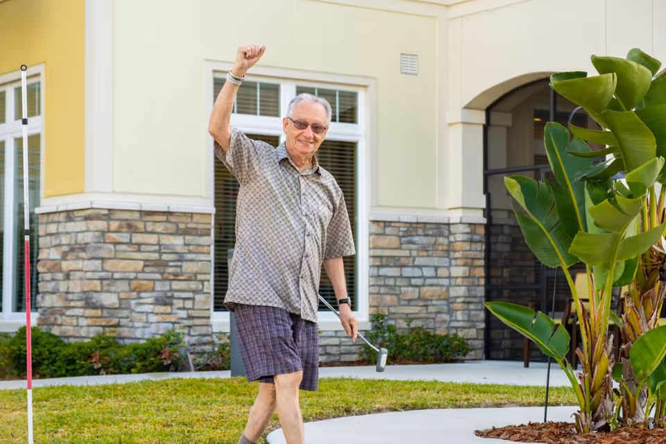 An elderly man wearing sunglasses, a patterned short-sleeve shirt, and shorts is outdoors on a lawn holding a mini golf putter and raising one arm in celebration. Behind him is a building with stone and yellow walls, windows, and some green plants.