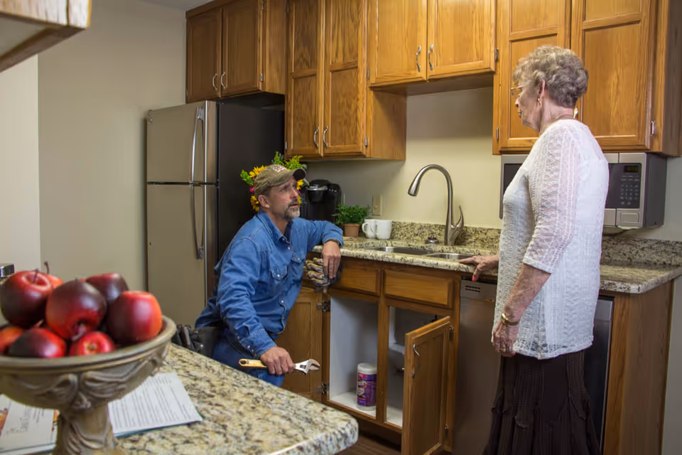 A man in a blue shirt and cap is kneeling and holding a wrench while repairing the cabinet under a kitchen sink. An elderly woman in a white lace top and brown skirt is standing nearby watching him. The kitchen has wooden cabinets, a granite countertop, a stainless steel refrigerator, a microwave, and a bowl of red apples on the counter.