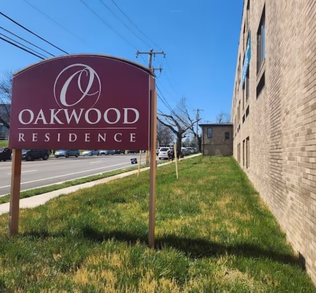 Oakwood Residence sign beside brick building on a grassy verge by the street.