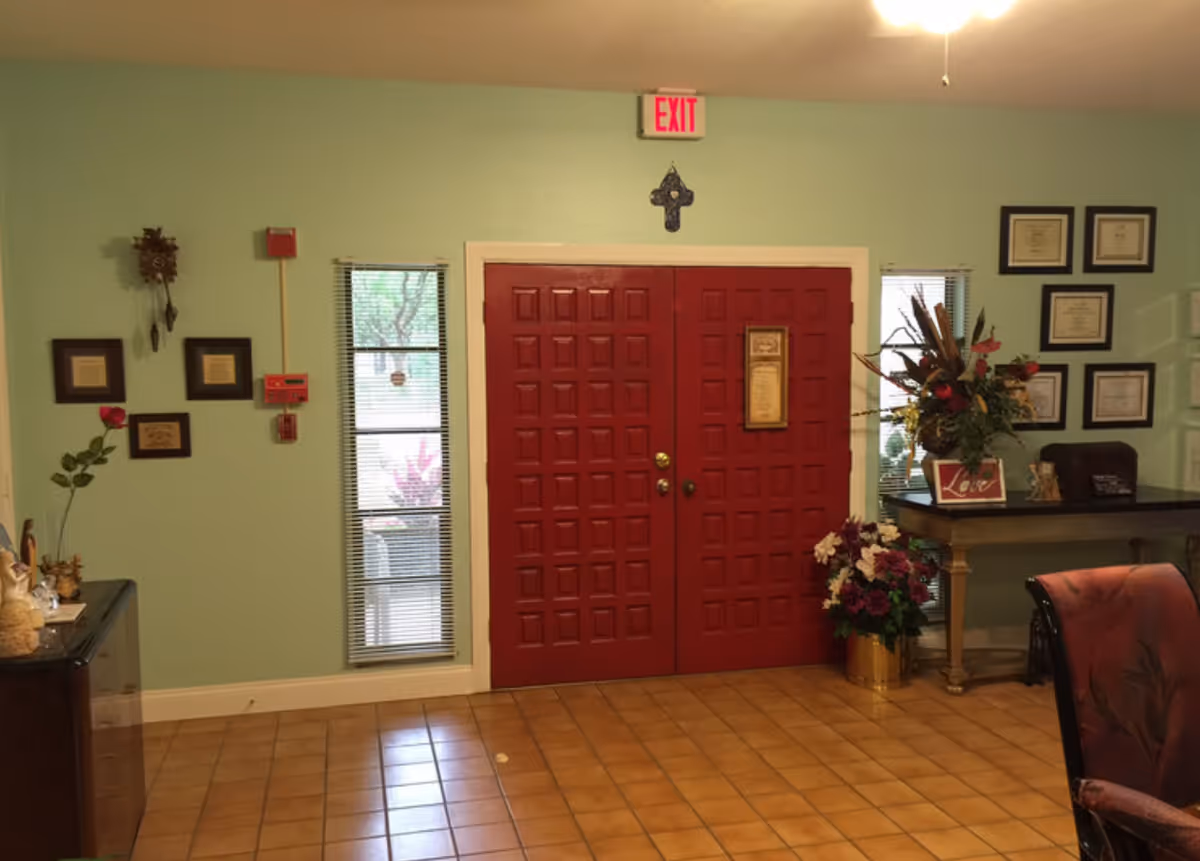 Interior view of an assisted living facility entrance area with double red doors, two narrow windows with blinds on either side, a green wall with framed certificates and decorations, a table with a flower arrangement and framed photo, and a tiled floor.