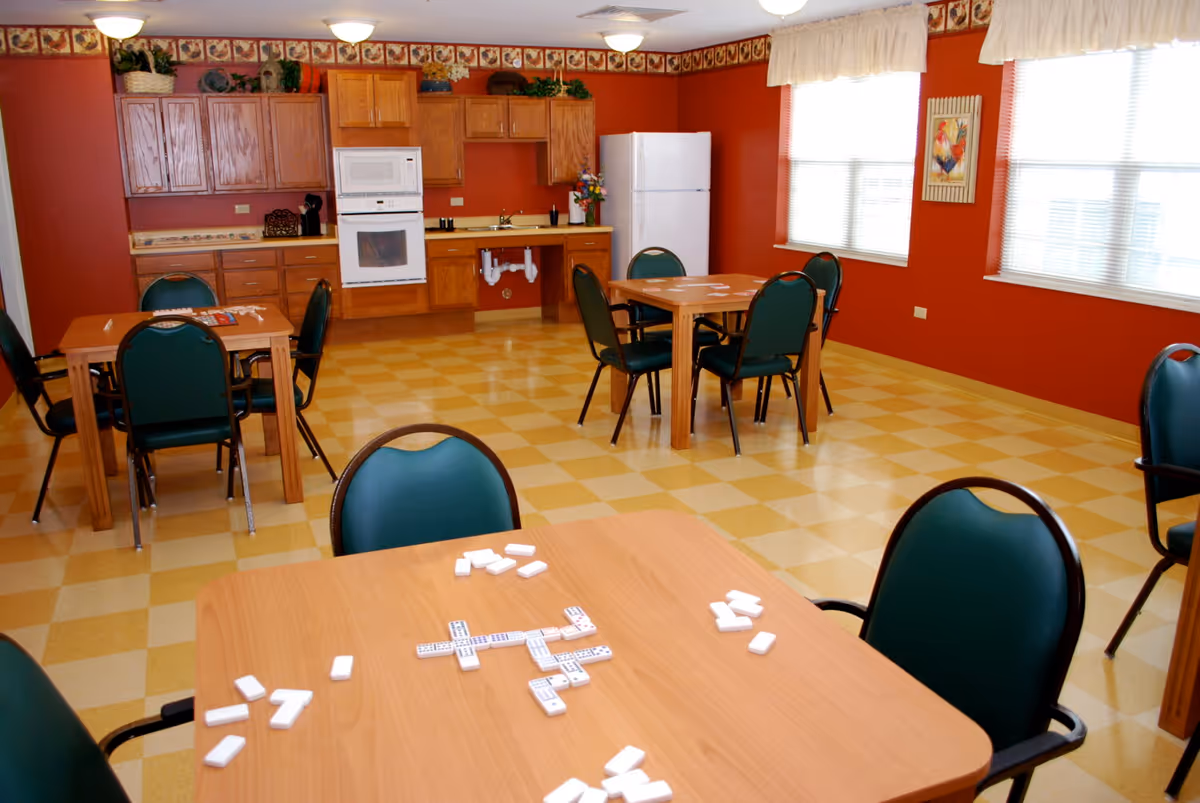 A bright room with orange walls and checkered yellow and beige flooring featuring several wooden tables with green cushioned chairs. Domino tiles are spread out on the tables. The room includes a kitchen area with wooden cabinets, a white refrigerator, a microwave, and an oven. Large windows with white curtains allow natural light to fill the space.