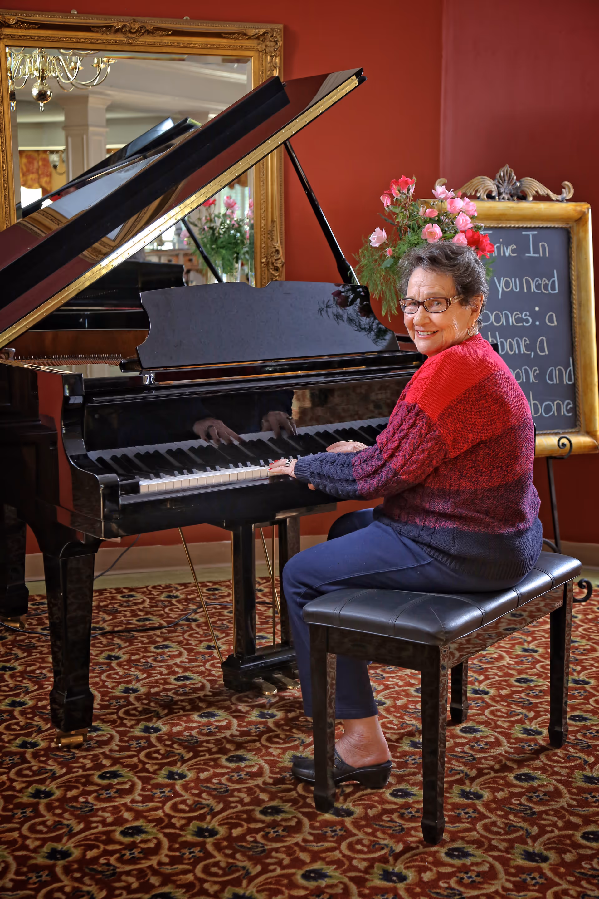 An elderly woman wearing glasses and a red and purple sweater is sitting on a black piano bench playing a grand piano in a room with a patterned carpet and red walls. Behind her, there is a large ornate gold-framed mirror and a chalkboard with flowers on top.