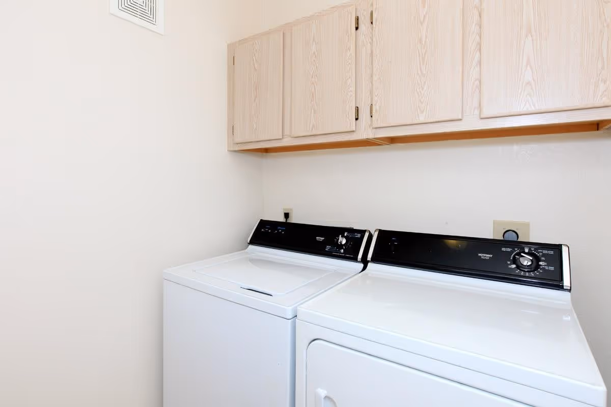 Laundry room with a white washing machine and dryer side by side under wooden cabinets mounted on a white wall.