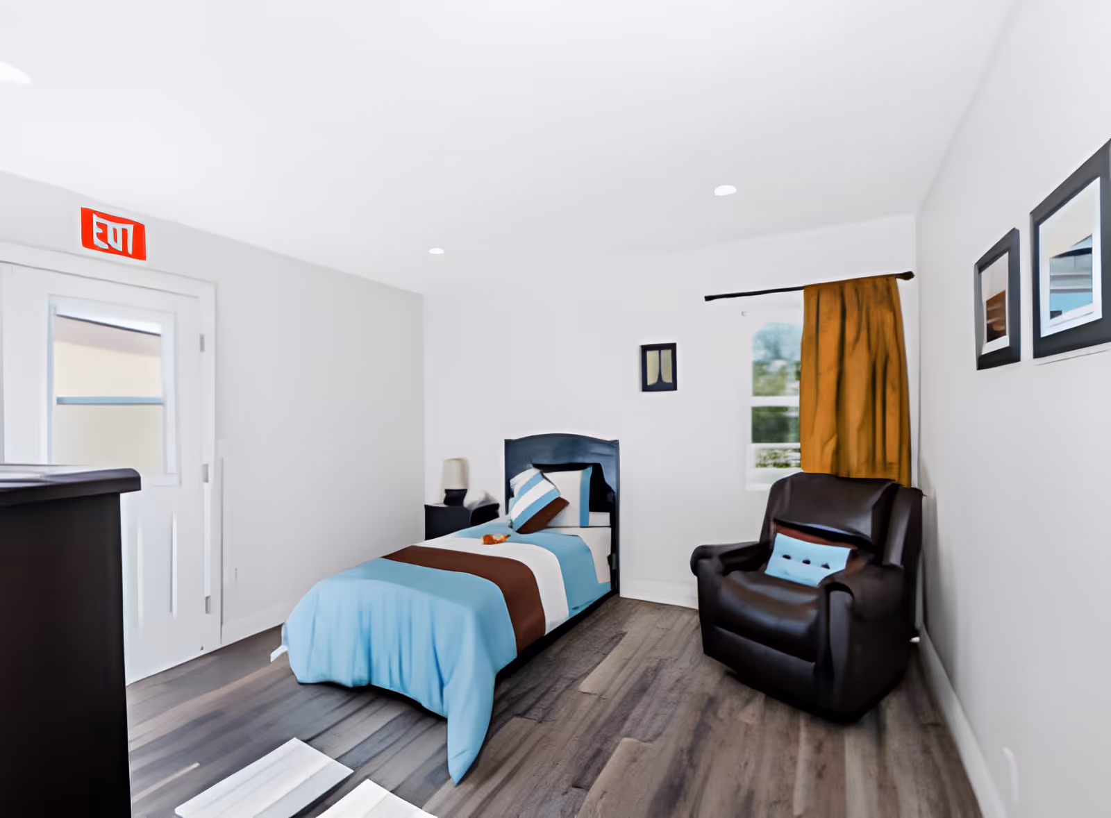 A simple bedroom with a single bed dressed in blue and brown bedding, a dark recliner chair, a window with a mustard curtain, and an exit sign above the door.
