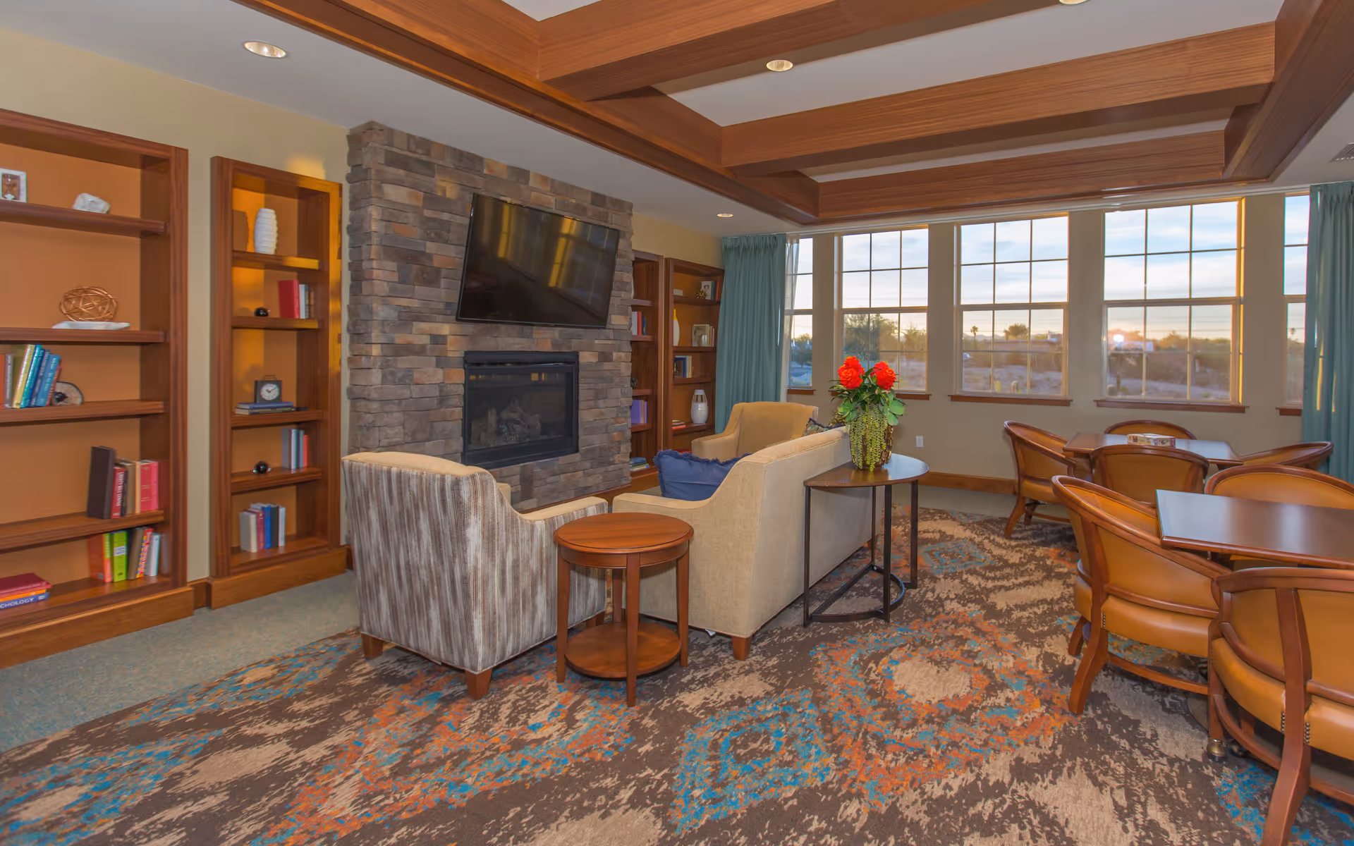 A cozy living room area with a stone fireplace and a mounted flat-screen TV above it. The room features a beige sofa, a striped armchair, wooden side tables, and built-in wooden bookshelves filled with books and decorative items. Large windows with teal curtains allow natural light to fill the space, and there are several wooden tables and chairs arranged near the windows. The ceiling has wooden beams, and the carpet has a colorful, patterned design.