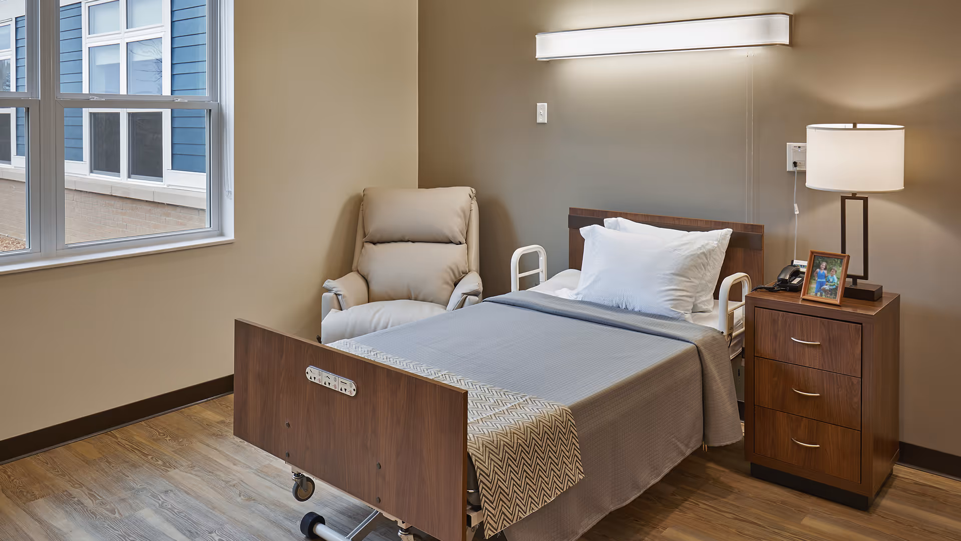 A neatly made single hospital-style bed with a gray blanket and white pillow in a room with beige walls and wood flooring. Next to the bed is a wooden nightstand with three drawers, a lamp, a telephone, and a framed photo. A beige recliner chair is positioned beside the bed near a large window showing the exterior of another building.