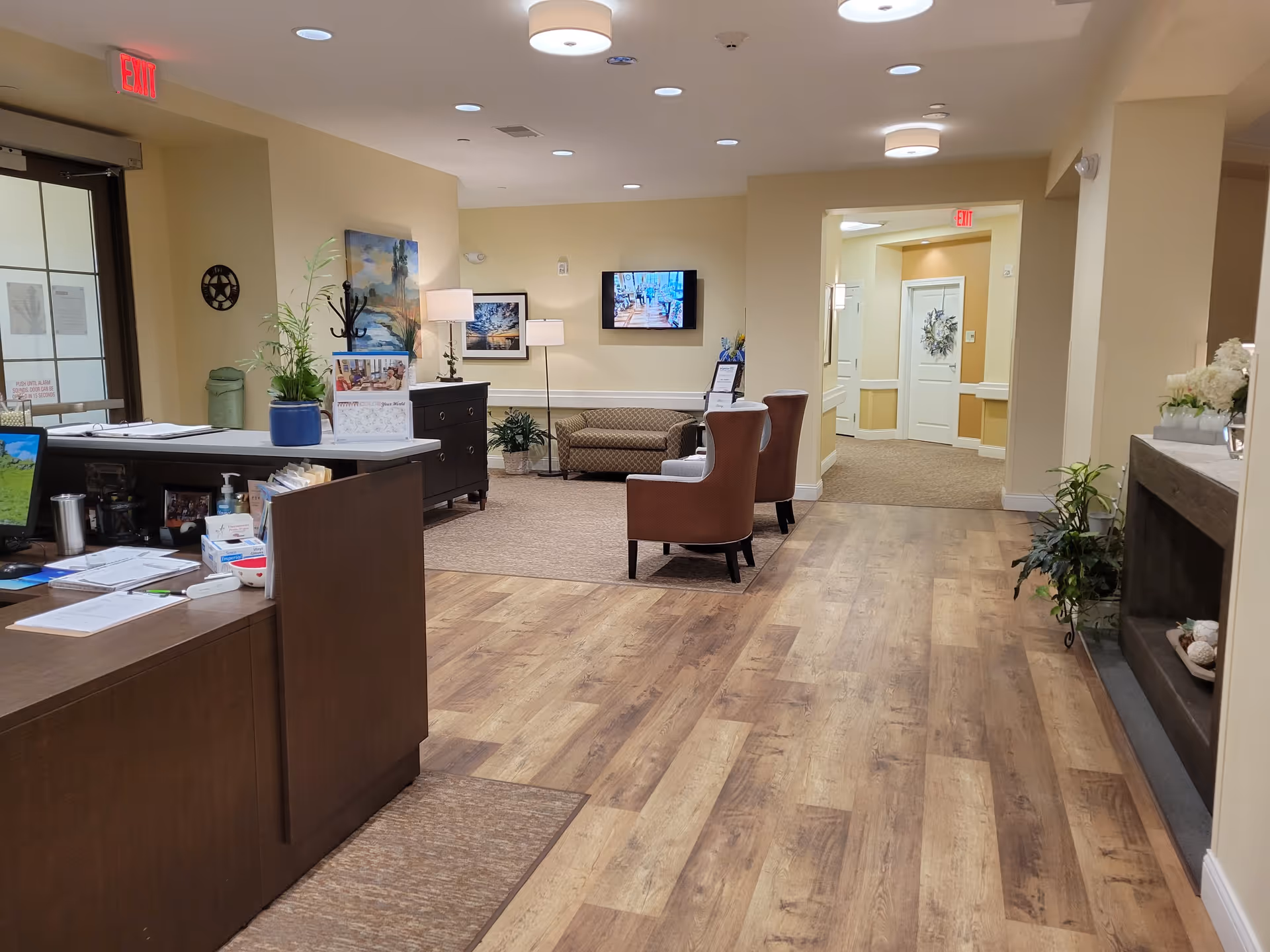 Interior view of a senior living facility reception area with a wooden desk on the left, a seating area with two brown chairs and a patterned loveseat, plants, framed artwork on the walls, a TV mounted on the far wall, and a hallway leading to white doors. The floor is a combination of wood and carpet, and the walls are painted beige.