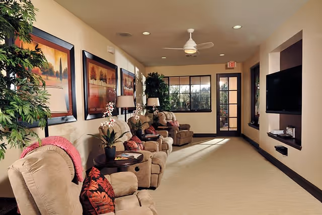 A cozy living room area with several beige recliner chairs arranged along the wall, each with a small side table and decorative pillows. The walls are adorned with framed landscape paintings, and there are potted plants adding greenery. A ceiling fan and recessed lighting are visible, and a flat-screen TV is mounted on the opposite wall. Large windows and a glass door let in natural light.