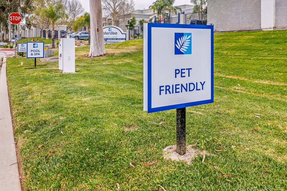 Outdoor view of Menifee Senior Living facility with green grass and several signs including a prominent 'PET FRIENDLY' sign in the foreground, a 'POOL & SPA' sign, a 'TOUR TODAY' sign, and a stop sign near the road. Buildings and trees are visible in the background.