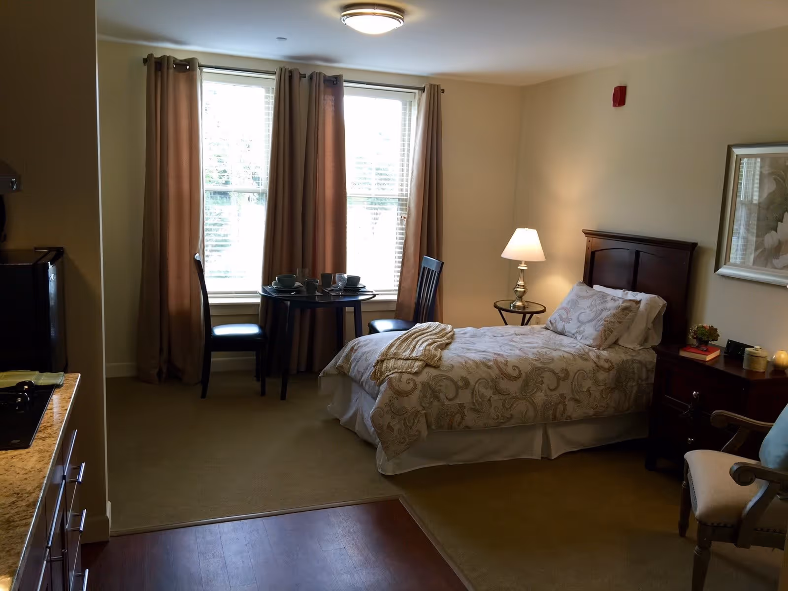 A cozy bedroom with a single bed covered in patterned bedding, a bedside table with a lamp, a small round dining table with two chairs near a window with brown curtains, and a kitchenette area with a stove and countertop on the left side.