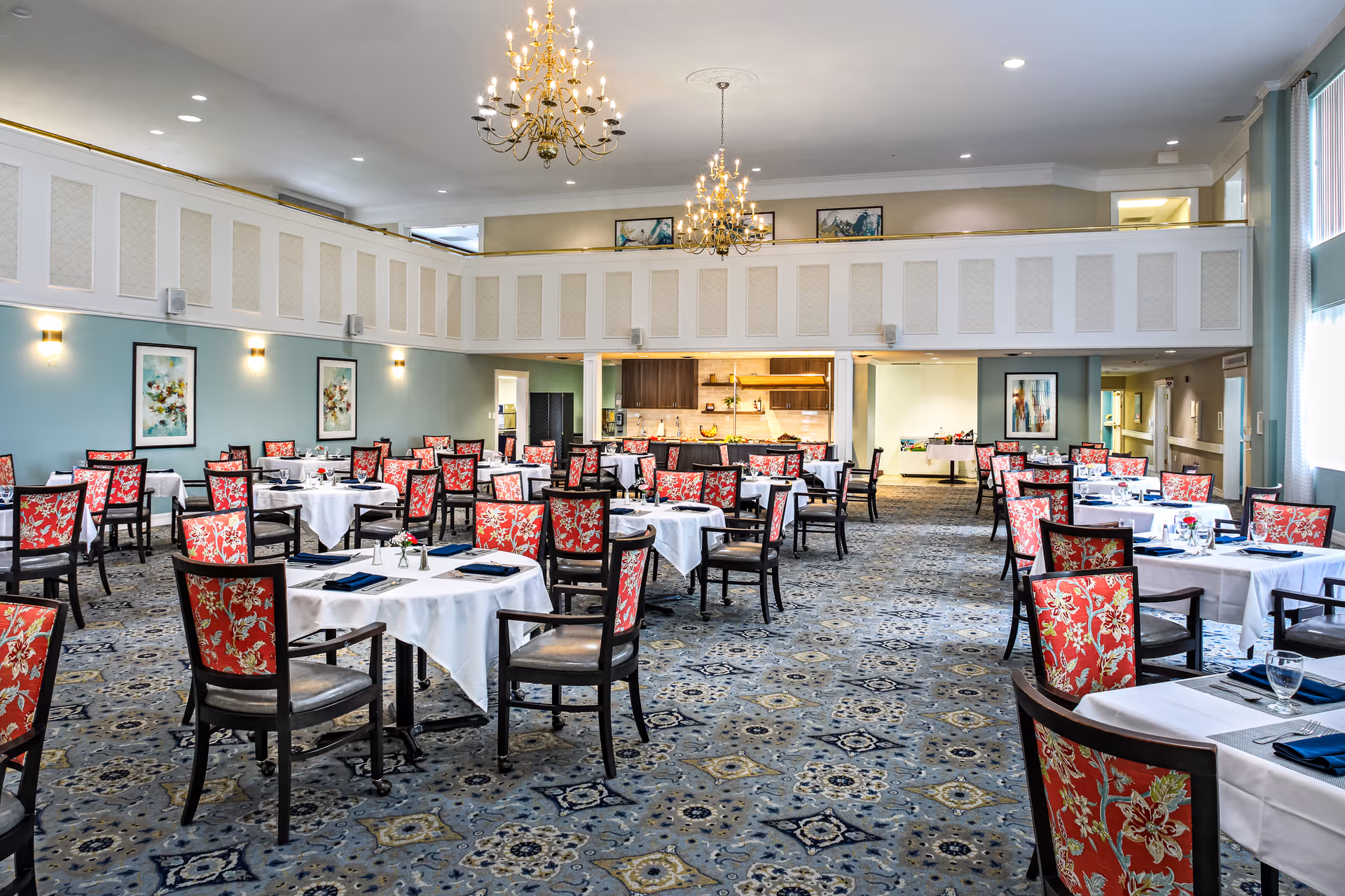 A spacious dining room with multiple round tables covered in white tablecloths, each surrounded by chairs with red floral upholstery. The room features a patterned carpet, light blue walls with framed artwork, two chandeliers hanging from the ceiling, and a kitchen area visible in the background.