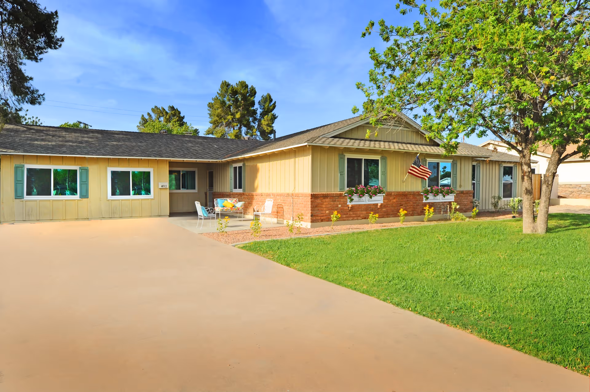 Single-story residential building with beige siding and brick accents, featuring green shutters and flower boxes under the windows. An American flag is mounted on the front wall. There is a concrete driveway and a green lawn with a tree on the right side. Outdoor seating with colorful cushions is placed near the entrance.