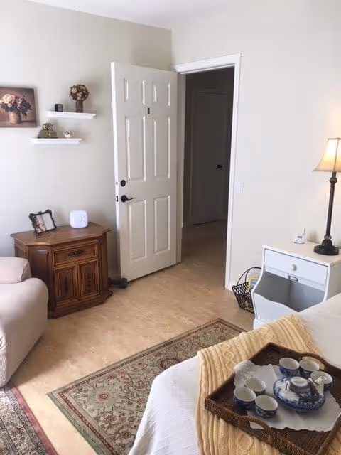 A cozy interior room with a partially open white door leading to a hallway. The room features a beige armchair, a wooden side table with decorative items, two white floating shelves with small decor, a white nightstand with a lamp, and a bed with a beige blanket and a tray holding a blue and white tea set. A patterned rug covers part of the floor.