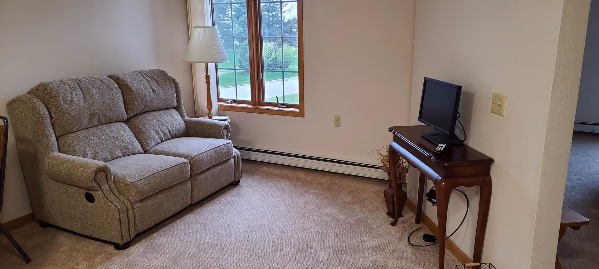 Small living room with a beige reclining loveseat by a window, a floor lamp, and a small wooden table holding a TV.