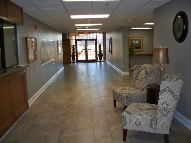 A long hallway with tiled flooring and gray walls leading to glass double doors at the far end. On the right side, there are two patterned upholstered chairs with a small table and lamp between them. On the left side, there is a wooden counter and a wall-mounted mail organizer. The hallway is well-lit with ceiling lights.
