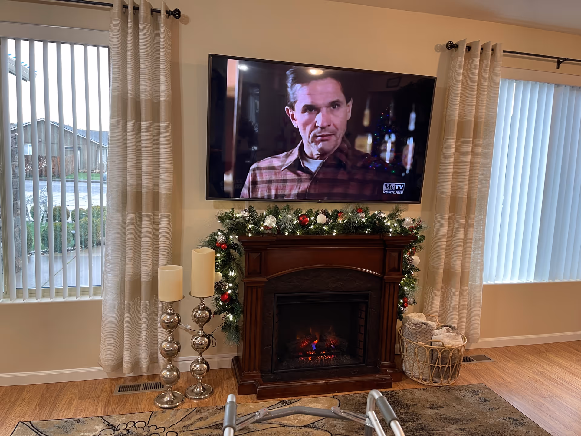 A cozy living room area featuring a wall-mounted flat-screen TV showing a man in a plaid shirt. Below the TV is a dark wooden electric fireplace decorated with a festive garland with lights and ornaments. To the left of the fireplace are two tall silver candle holders with white candles. To the right is a basket filled with rolled blankets. The room has light-colored walls, two windows with vertical blinds and beige curtains, and a wooden floor with a patterned rug partially visible. A walker is also partially visible in the foreground.
