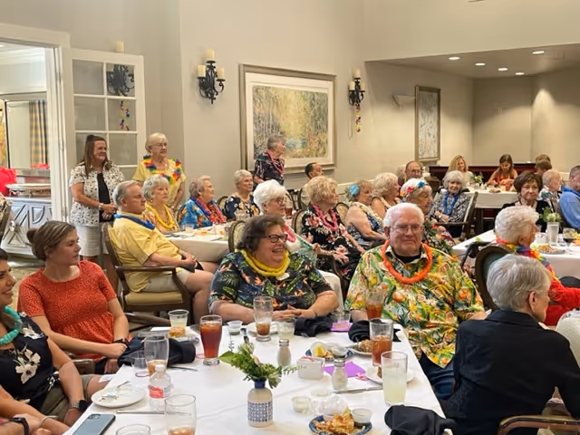 A group of elderly people seated around tables in a dining room, enjoying a social event with drinks and food. Some attendees are wearing colorful leis and festive clothing. The room has light-colored walls with framed artwork and wall sconces.