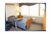 A small, simple bedroom in a nursing facility with a single bed covered in a blue quilt, a bedside table with a lamp, a window with curtains, and a small overbed table positioned next to the bed.