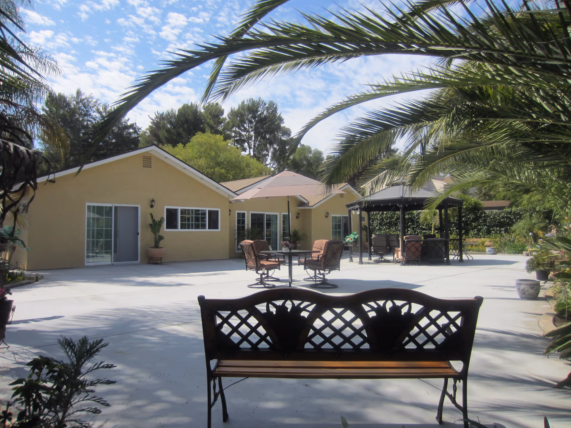 Outdoor courtyard with a bench in the foreground, patio seating and a gazebo in front of a single-story yellow building framed by palm trees.