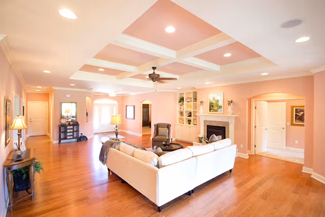 A spacious living room with light pink walls and a coffered ceiling with recessed lighting and a ceiling fan. The room features a large white sectional sofa, a single armchair, a round coffee table, a fireplace with built-in shelves on either side, and wooden flooring. There are multiple doorways leading to other rooms and a hallway with additional furniture and lamps.