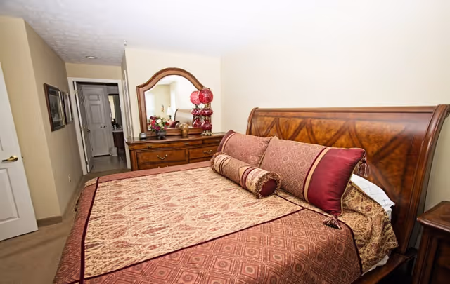 A bedroom with a large wooden bed featuring an ornate headboard and patterned bedding in shades of burgundy and beige. There are several decorative pillows on the bed. Against the wall is a wooden dresser with a large mirror, adorned with a red lamp and a floral arrangement. The room has beige walls and carpet, with a hallway and door visible in the background.