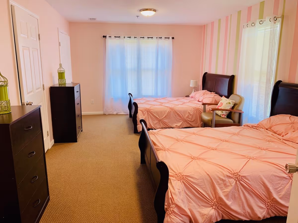 A bright bedroom with two twin beds dressed in pink comforters, dark wood dressers, an armchair, and a striped accent wall by curtained windows.