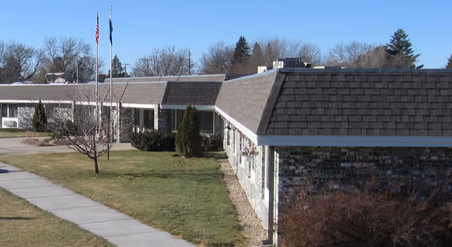 Exterior view of a single-story senior living facility building with a shingled roof, several windows, and a well-maintained lawn with a sidewalk leading to the entrance. Two flagpoles with flags are visible near the entrance, and leafless trees and shrubs surround the building under a clear blue sky.