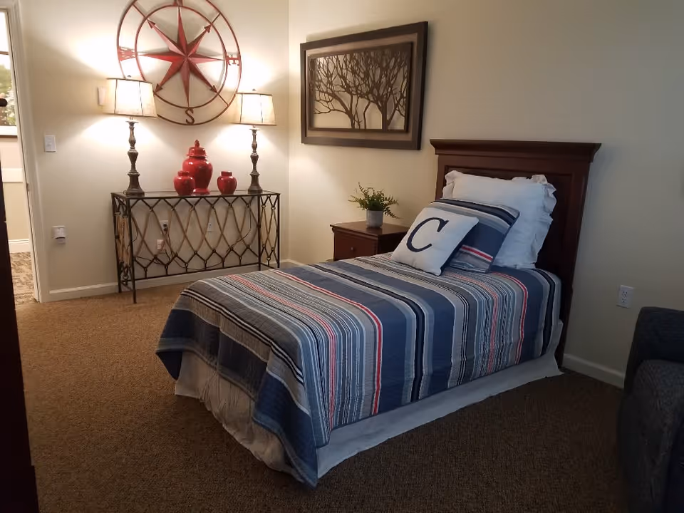 A cozy bedroom with a single bed covered in a striped blue, gray, and red quilt. The bed has a white pillow and a decorative pillow with the letter 'C'. Next to the bed is a wooden nightstand with a small potted plant. On the wall above the nightstand is a framed artwork of tree branches. Across from the bed is a decorative metal console table with three red vases and two table lamps with beige shades. Above the console table is a large wall decoration resembling a compass rose. The room has beige walls and carpeted floor.