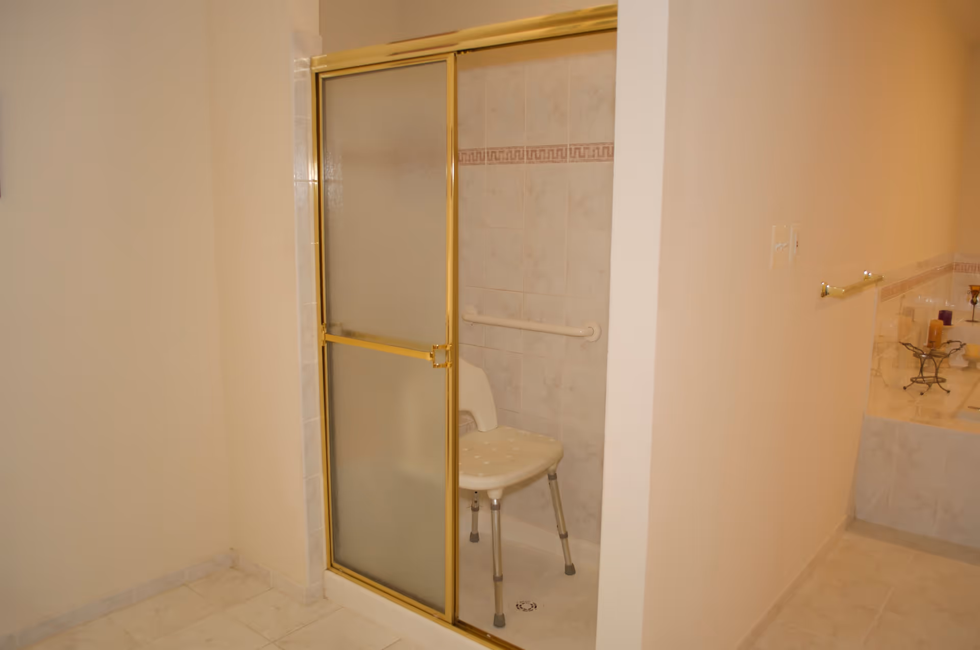 Bathroom with a shower area featuring a frosted glass sliding door with a gold frame. Inside the shower is a white plastic shower chair with metal legs and a grab bar mounted on the tiled wall. Adjacent to the shower is a bathtub with candles placed on the edge.
