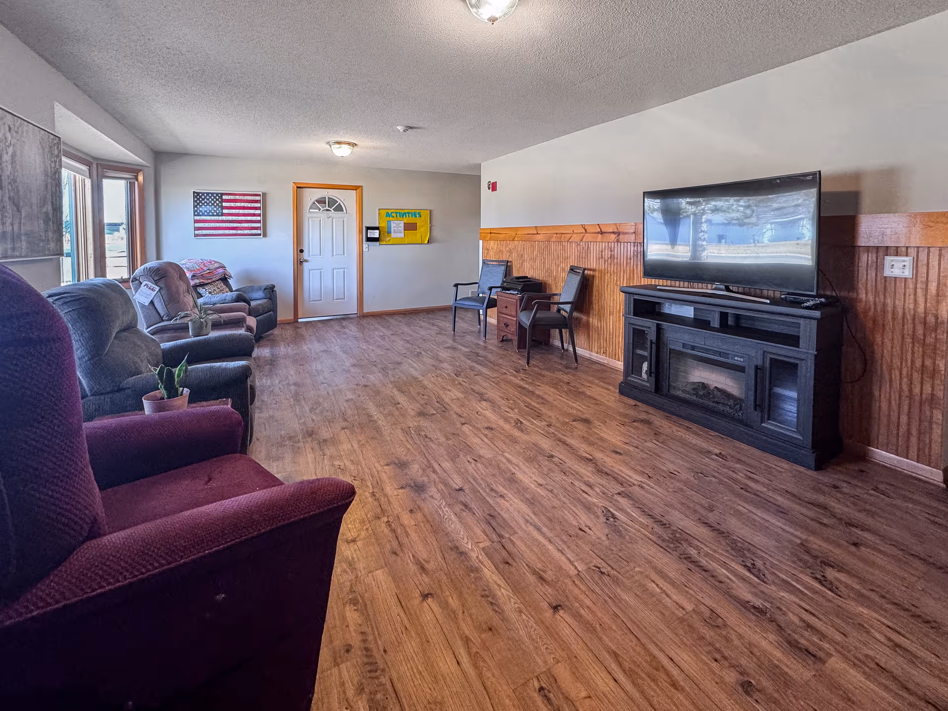 A spacious living room with wooden flooring and a row of armchairs along the left wall. There is a large flat-screen TV on a black stand with a built-in electric fireplace on the right side. The room has a white door with a window, an American flag hanging on the wall, and a yellow bulletin board labeled 'ACTIVITIES'. The walls are painted light gray with wooden paneling halfway up on the right side.