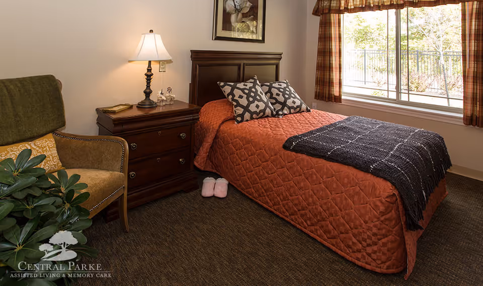 A cozy bedroom with a single bed covered in a rust-colored quilt and two patterned pillows. A dark wooden nightstand with a lamp and decorative items sits beside the bed. There is a comfortable armchair with a green throw and a yellow cushion next to a leafy green plant. A large window with plaid curtains lets in natural light, and a framed picture hangs above the bed.