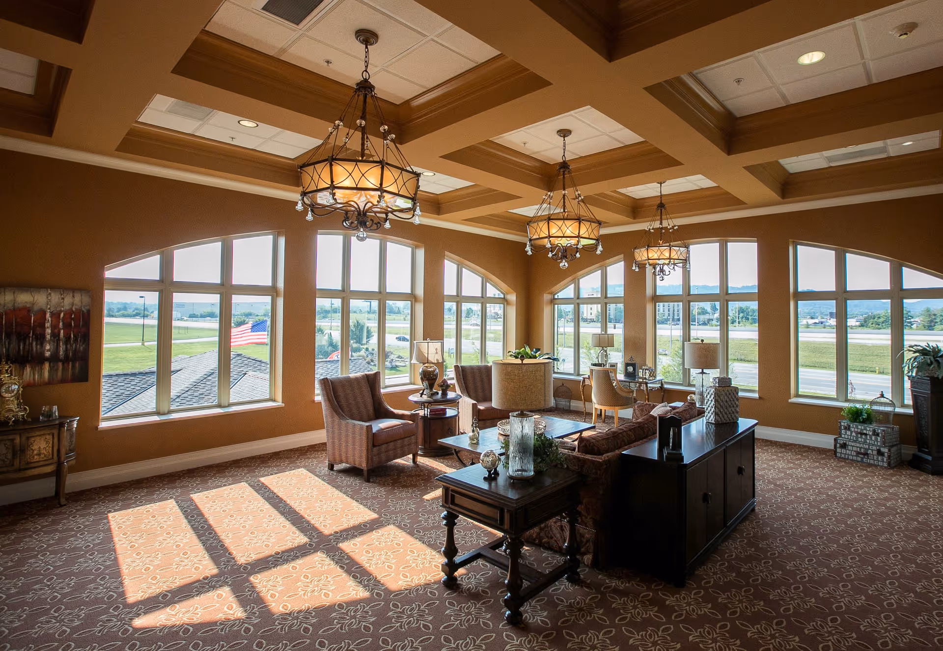A spacious, well-lit living room area with large arched windows allowing natural light to fill the space. The room features patterned carpet, multiple seating options including armchairs and sofas, wooden side tables with decorative items, and elegant hanging chandeliers. Outside the windows, a view of a green lawn, a flagpole with an American flag, and distant buildings is visible.